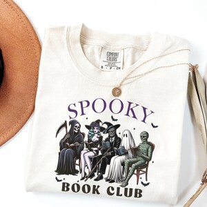 May include: Cream-colored t-shirt with the words "SPOOKY BOOK CLUB" in purple. The graphic features cartoon figures of the Grim Reaper, a witch, a skeleton, a ghost, and a mummy reading books. A gold necklace and a brown hat are also visible.