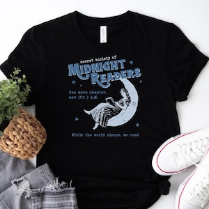 May include: Black t-shirt featuring the text "secret society of MIDNIGHT READERS" with a woman reading on a crescent moon. The shirt also includes the phrases "One more chapter, now it's 3 A.M." and "While the world sleeps, we read."