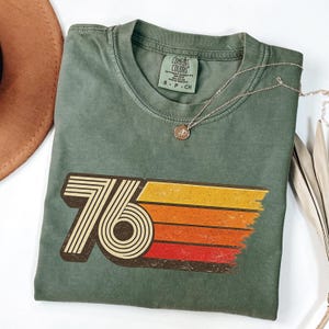May include: A sage green t-shirt with a retro "76" graphic in brown, yellow, orange, and red. The shirt has a crew neck and short sleeves. A silver necklace with a small pendant is draped across the shirt.
