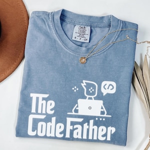 May include: A blue short-sleeved t-shirt with a white graphic print featuring a person at a laptop and the text The Code Father. The shirt has a Comfort Colors label at the neck and is displayed with a gold-toned necklace featuring a small round pendant.