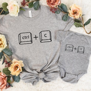 May include: A gray t-shirt and baby onesie set. The adult t-shirt has the text "ctrl + C" printed in black. The baby onesie has the text "ctrl + V" printed in black. The set is surrounded by flowers.