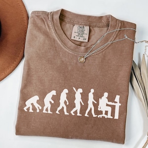 May include: A brown short-sleeved t-shirt featuring a white graphic print showing the evolution of humans, ending with a figure sitting at a computer desk. The shirt has a Comfort Colors brand label at the neck and is paired with a silver-toned necklace.