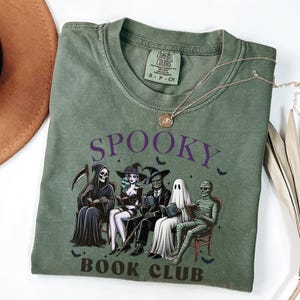 May include: Olive green t-shirt with the words "Spooky Book Club" and a graphic of Halloween characters reading books. The shirt has a crew neck and short sleeves. A gold necklace is draped across the shirt.