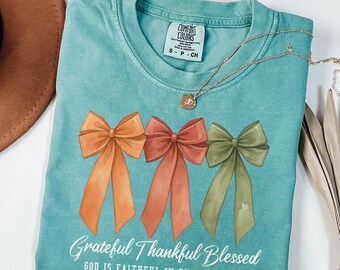 May include: A turquoise Comfort Colors t-shirt featuring three watercolor bows in orange, red, and green. The shirt has the text "Grateful Thankful Blessed" and "GOD IS FAITHFUL IN EVERY SEASON" in white script. A gold necklace is draped across the shirt.