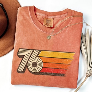 May include: A coral-colored t-shirt with a retro "76" graphic in brown and a striped orange, yellow, and red design. A gold necklace with a pendant rests on the shirt. The shirt has a Comfort Colors tag.