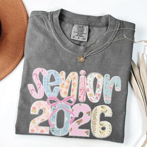 May include: A gray Comfort Colors t-shirt with the word "Senior 2026" in colorful floral and patterned fabric. A silver necklace with a pendant is draped across the shirt.