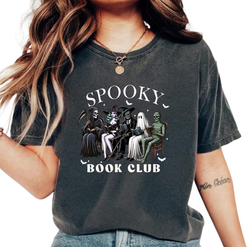 May include: Dark gray t-shirt featuring a graphic of a spooky book club with the text "SPOOKY BOOK CLUB". The graphic includes a Grim Reaper, a witch, a vampire, a ghost, and a mummy. Bats are scattered around the text and characters.