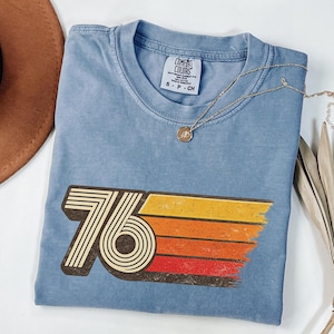 May include: A blue t-shirt with a retro "76" graphic in brown, yellow, orange, and red. The shirt has a crew neck and short sleeves. A gold necklace with a pendant is draped across the shirt.
