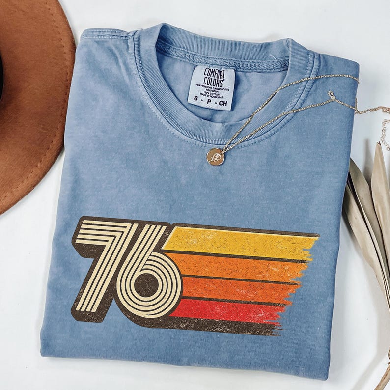 May include: A blue t-shirt with a retro "76" graphic in brown, yellow, orange, and red. The shirt has a crew neck and short sleeves. A gold necklace with a pendant is draped across the shirt.