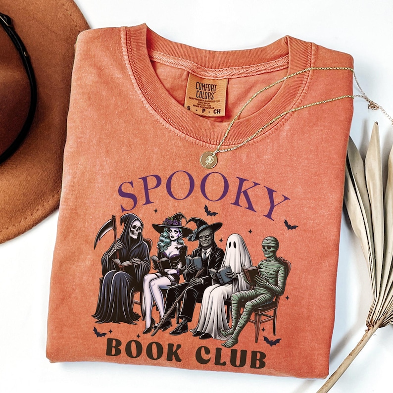 May include: Orange Comfort Colors t-shirt with a crew neck and the words "Spooky Book Club" in purple. The graphic features a Grim Reaper, a witch, a zombie, a ghost, and a mummy reading books. A gold necklace with a pendant is draped on the shirt.