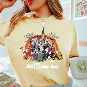 May include: A light yellow t-shirt featuring a Halloween-themed graphic of Disney characters in costume in front of a castle. The graphic includes Mickey and Minnie Mouse, Donald Duck, Goofy, and Pluto. The text "Trick or Treat" is on a tombstone.
