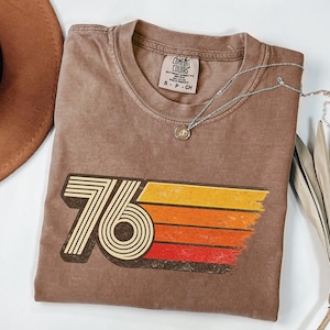 May include: A brown Comfort Colors t-shirt with a retro "76" graphic in white, yellow, orange, and red. A silver necklace with a small pendant rests on the shirt. The shirt is a small size.