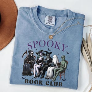 May include: A light blue t-shirt with the words "SPOOKY BOOK CLUB" in purple. The graphic features cartoon figures of the Grim Reaper, a witch, a zombie, a ghost, and a mummy reading books. A gold necklace is draped across the shirt.