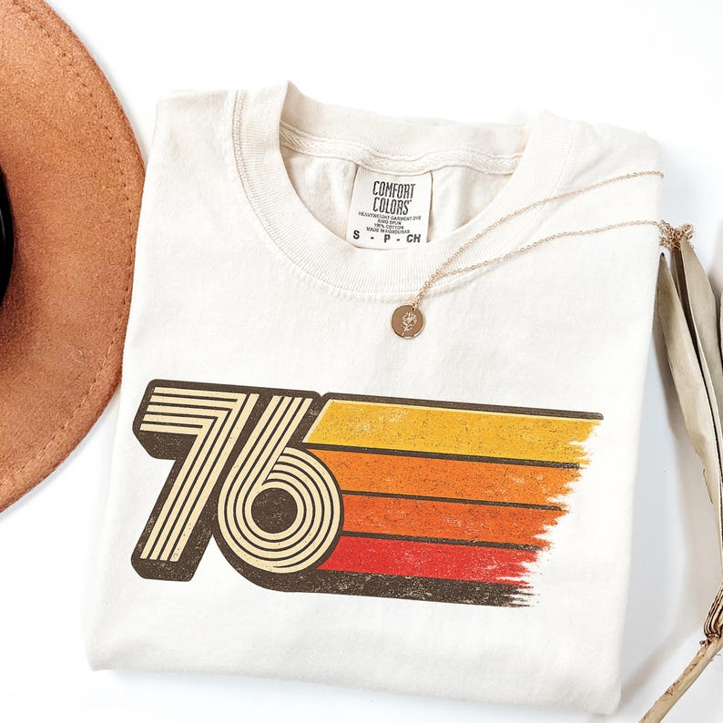 May include: Cream-colored t-shirt with a retro "76" graphic in black, orange, and yellow. A gold necklace with a pendant and a brown hat are next to the shirt. The shirt is made by Comfort Colors.