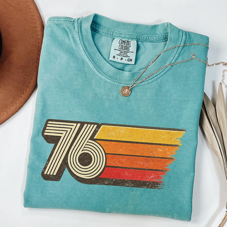 May include: A turquoise Comfort Colors t-shirt with a retro "76" graphic in brown, yellow, orange, and red. A gold necklace with a pendant rests on the shirt. A brown hat is visible in the upper left corner.