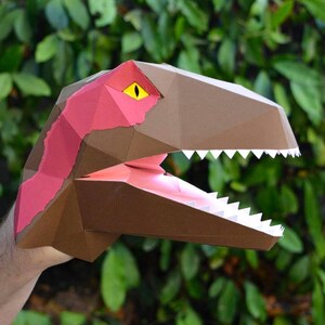 May include: A brown and pink papercraft dinosaur head with a yellow eye and white teeth. The dinosaur head is open-mouthed and appears to be roaring.
