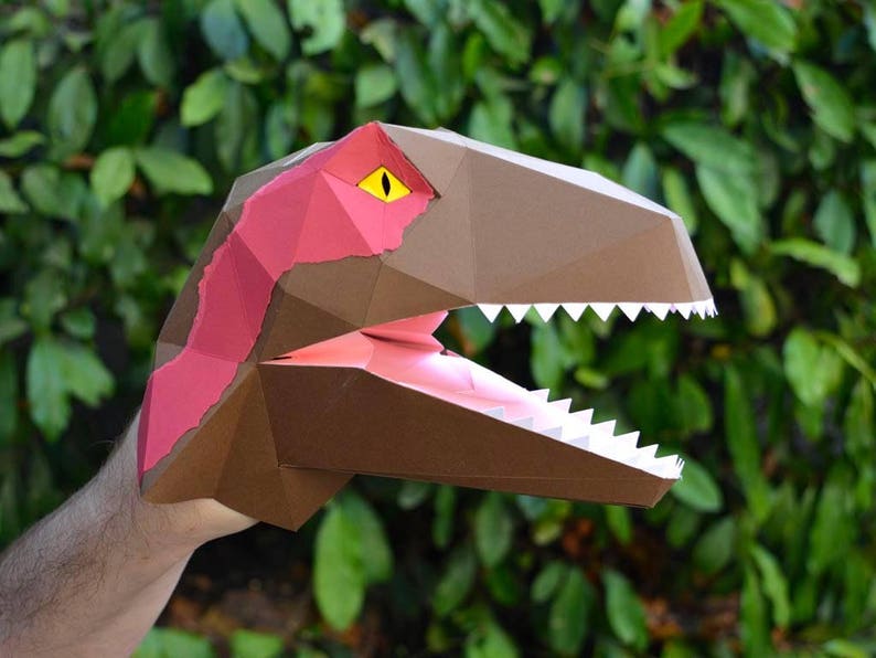 May include: A brown and pink papercraft dinosaur head with a yellow eye and white teeth. The dinosaur head is open-mouthed and appears to be roaring.