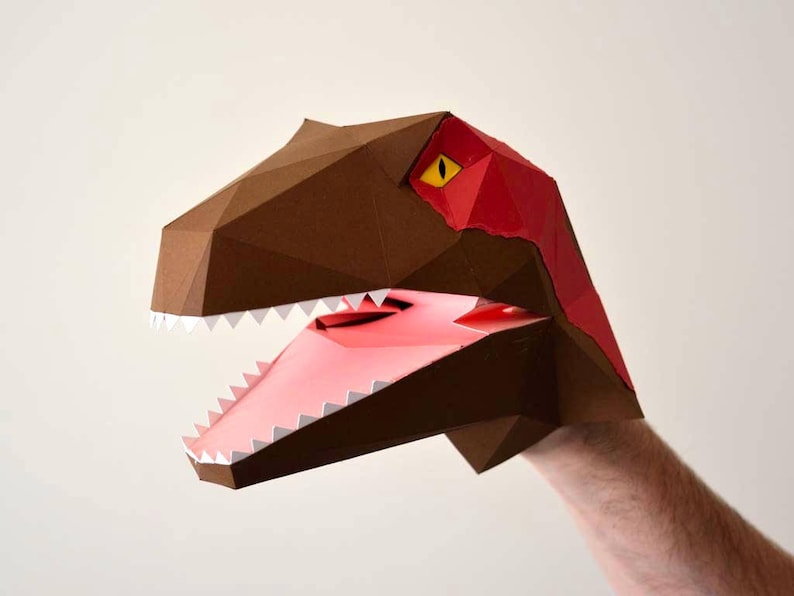 May include: A brown paper dinosaur puppet with a red and yellow eye and a pink mouth with white teeth.