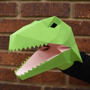 May include: A green paper dinosaur puppet with a pink open mouth and sharp teeth. The puppet is held by a hand wearing a black sleeve.