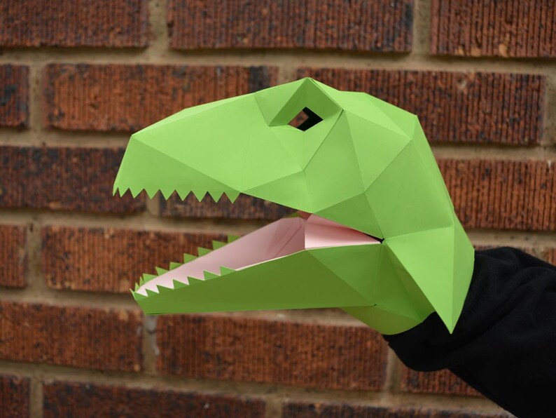 May include: Green paper dinosaur puppet with a pink mouth, ready to be used for storytelling or puppet shows.