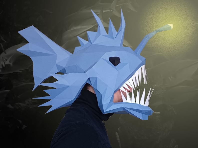 May include: A blue paper mache anglerfish mask with sharp teeth and a glowing lure. The mask is worn by a person, but the person's face is not visible.