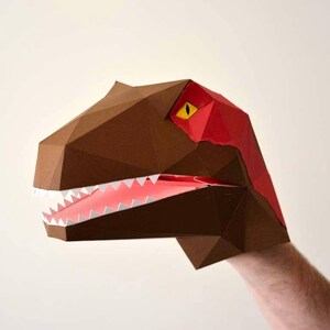 May include: A brown papercraft dinosaur puppet with a red and yellow eye and a red mouth. The puppet is being held by a hand.