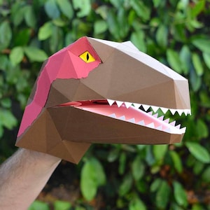 May include: A brown papercraft dinosaur puppet with a pink mouth and yellow eye. The puppet is held by a hand.