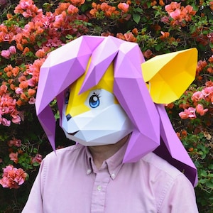Peut inclure: Une personne portant un masque en papier rose et jaune avec un motif de renard. Le masque a une crinière rose et des oreilles jaunes. La personne est debout devant un buisson avec des fleurs roses.