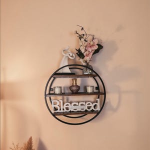 May include: A round, black metal wall shelf with three wooden shelves. The shelf holds a small vase, two candles, and a decorative figurine. The word "Blessed" is displayed in white letters on the bottom shelf. Pink flowers are in a glass vase.