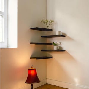 May include: A corner wall shelf unit with three dark brown shelves. The shelves hold a white mug and small plants. A red table lamp with a black base sits on the wooden floor. The room has white walls and a window.