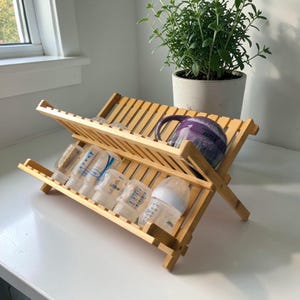 May include: A natural bamboo dish rack holds baby bottles and a purple mug. The rack is angled, with two tiers for drying items. A potted green plant sits in the background near a window.