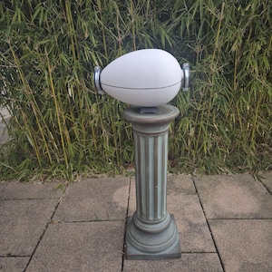 Pode incluir: Um objeto branco em forma de ovo com detalhes prateados está em cima de um pedestal verde. O pedestal tem uma coluna canelada e uma base quadrada. O objeto está na frente de um fundo de bambu verde.