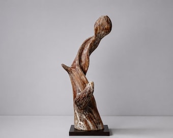 May include: A vertical sculpture crafted from a piece of weathered driftwood with a natural brown and white grain pattern. The wood features a twisted, organic shape that curves upward and is mounted on a square, dark brown wooden base.