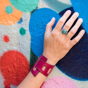 May include: A close-up of a woman's wrist wearing a pink leather cuff bracelet with a unique cut-out design. The bracelet is on her left wrist, and she is wearing a ring with a turquoise stone on her right hand.