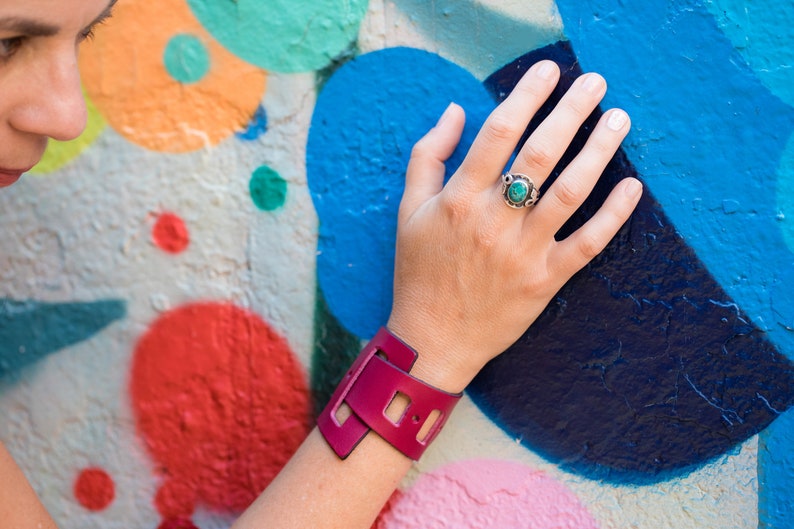 May include: A close-up of a woman's wrist wearing a pink leather cuff bracelet with a unique cut-out design. The bracelet is on her left wrist, and she is wearing a ring with a turquoise stone on her right hand.