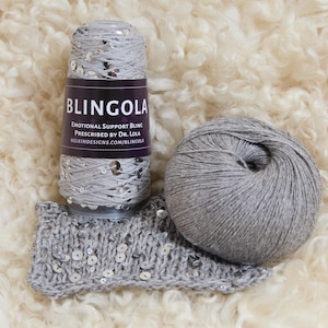 Puede incluir: Un ovillo de hilo gris con lentejuelas, una bola de hilo gris y una muestra tejida con lentejuelas. El hilo está etiquetado "BLINGOLA" con el texto "EMOTIONAL SUPPORT BLING PRESCRIBED BY DR. LOLA NELKINDESIGNS.COM/BLINGOLA".