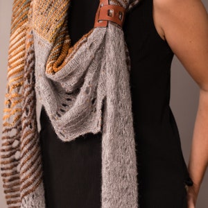 May include: A gray and brown knitted scarf with a leather clasp. The scarf is draped around a person's neck.