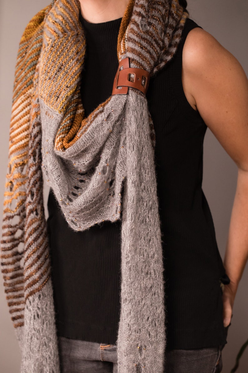 May include: A gray and brown knitted scarf with a leather clasp. The scarf is draped around a person's neck.