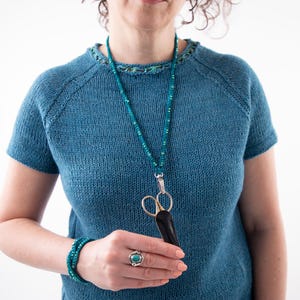 Pu&ograve; includere: Un top blu lavorato a maglia con maniche corte e girocollo. Una collana di perline turchese con un ciondolo a forbice argentato pende dal collo. Sono visibili anche un braccialetto turchese e un anello con una pietra turchese.