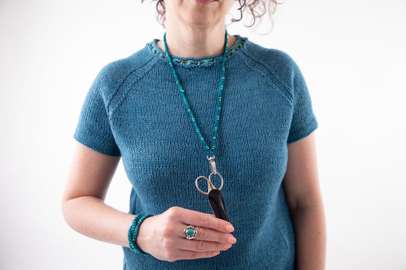 Pu&ograve; includere: Un top blu lavorato a maglia con maniche corte e girocollo. Una collana di perline turchese con un ciondolo a forbice argentato pende dal collo. Sono visibili anche un braccialetto turchese e un anello con una pietra turchese.