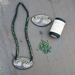 May include: A handmade necklace with a tree pendant, featuring a green beaded chain. The image also shows a matching pendant, beads, thread, and a spool of green twine. The pendants have a tree design with blue and brown accents.