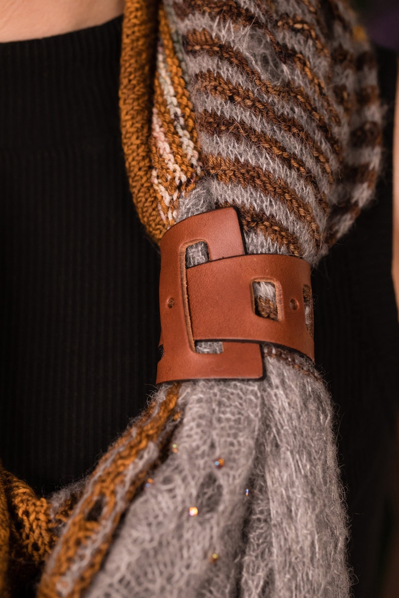 May include: A brown leather scarf holder with a unique design. The holder is attached to a gray and brown striped scarf.