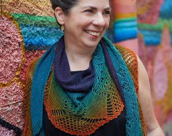 May include: A hand-knitted triangular shawl featuring a vibrant gradient of colours transitioning from deep purple to teal, orange, and yellow. The shawl has an intricate lace pattern along the edges, accented with small beads along the bottom border.