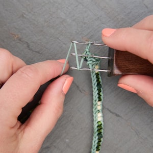 Pu&ograve; includere: Primo piano di un braccialetto verde e con perline in fase di realizzazione. Il braccialetto viene tessuto con un piccolo telaio e un manico in legno. Il braccialetto &egrave; verde con perline bianche e verdi. Lo sfondo &egrave; una superficie grigia.