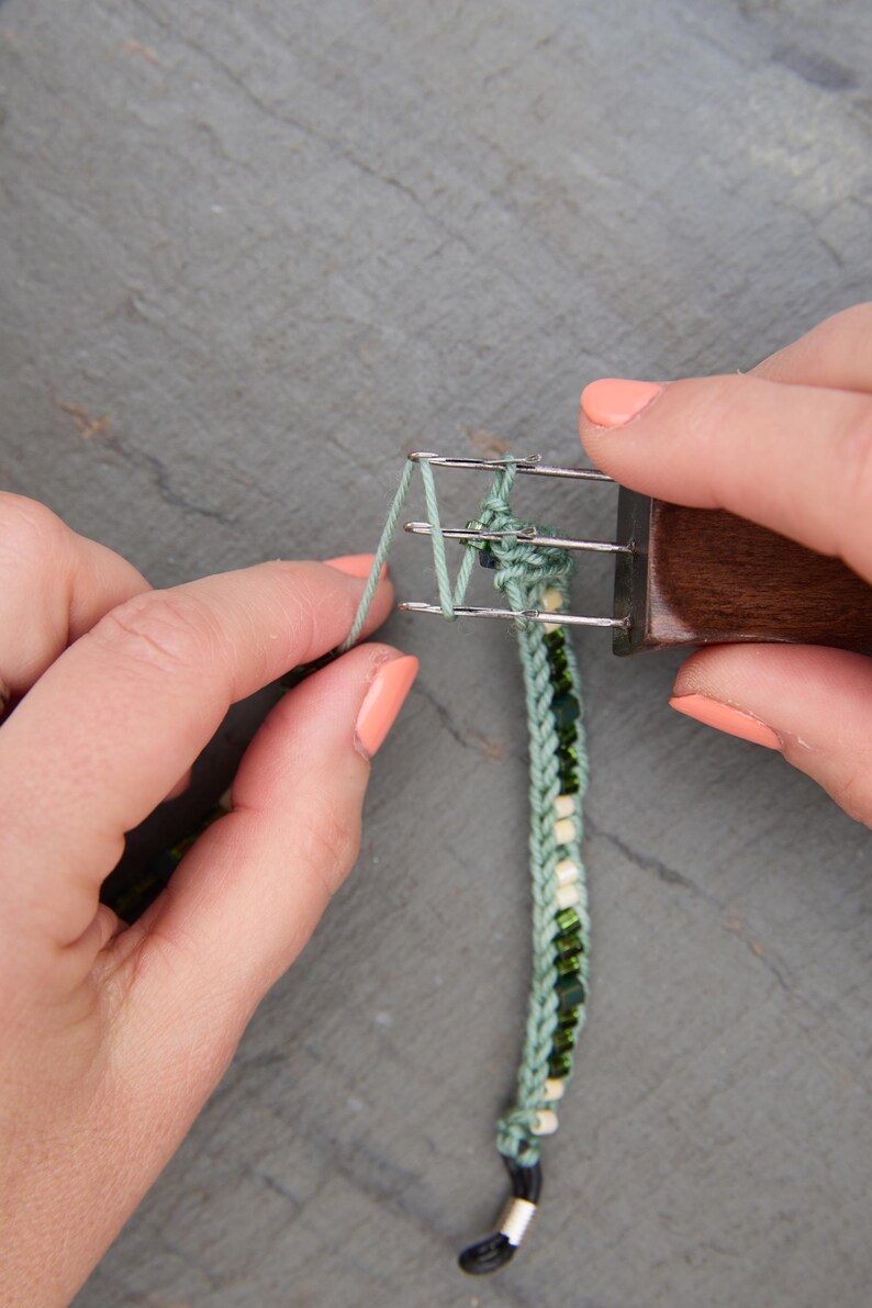 Pu&ograve; includere: Primo piano di un braccialetto verde e con perline in fase di realizzazione. Il braccialetto viene tessuto con un piccolo telaio e un manico in legno. Il braccialetto &egrave; verde con perline bianche e verdi. Lo sfondo &egrave; una superficie grigia.