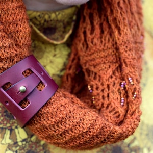 May include: A close-up of a brown knitted scarf with a burgundy leather bracelet. The bracelet has a buckle and a cut-out design.