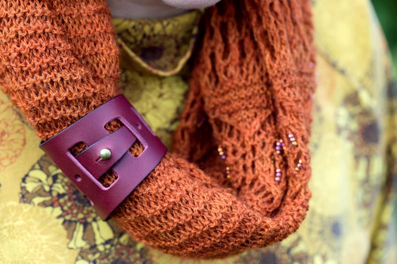 May include: A close-up of a brown knitted scarf with a burgundy leather bracelet. The bracelet has a buckle and a cut-out design.