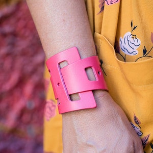 May include: A pink leather cuff bracelet with a geometric design. The bracelet is wide and has a cutout design.
