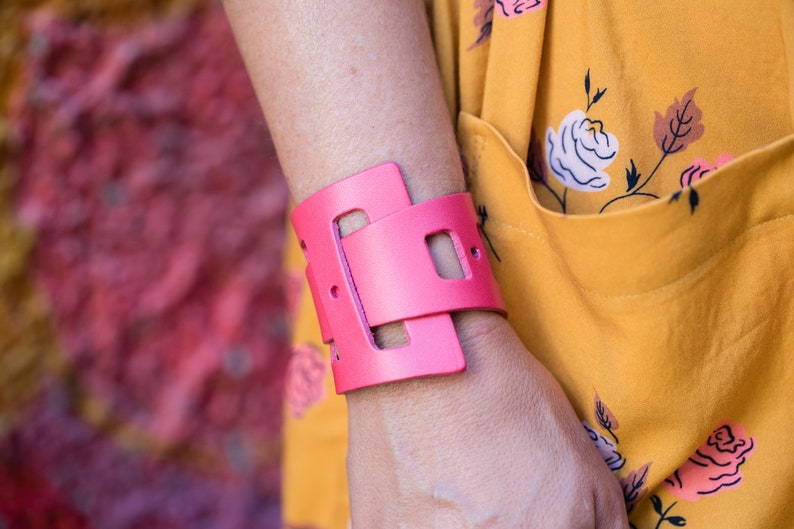 May include: A pink leather cuff bracelet with a geometric design. The bracelet is wide and has a cutout design.