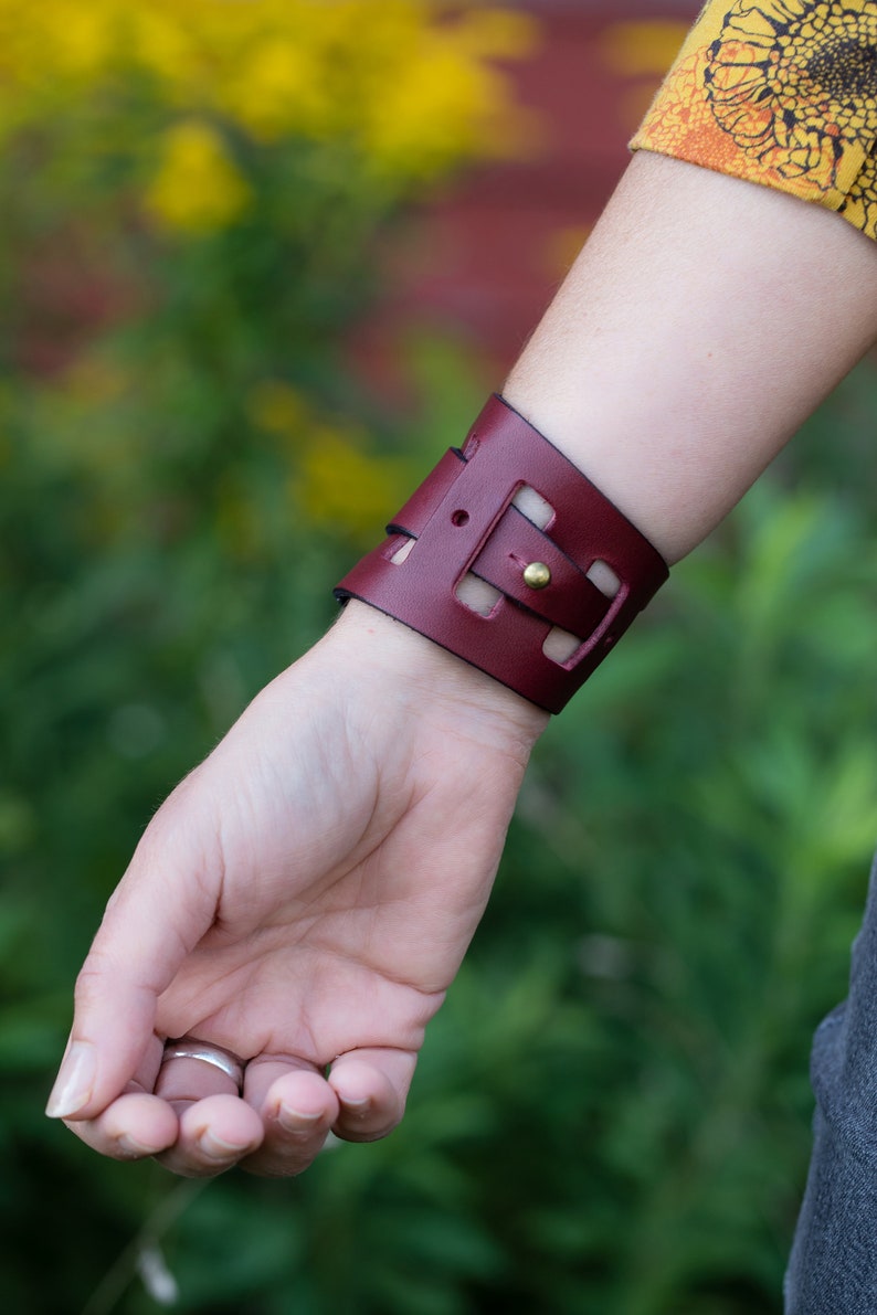 May include: A burgundy leather cuff bracelet with a cut-out design and a brass buckle.